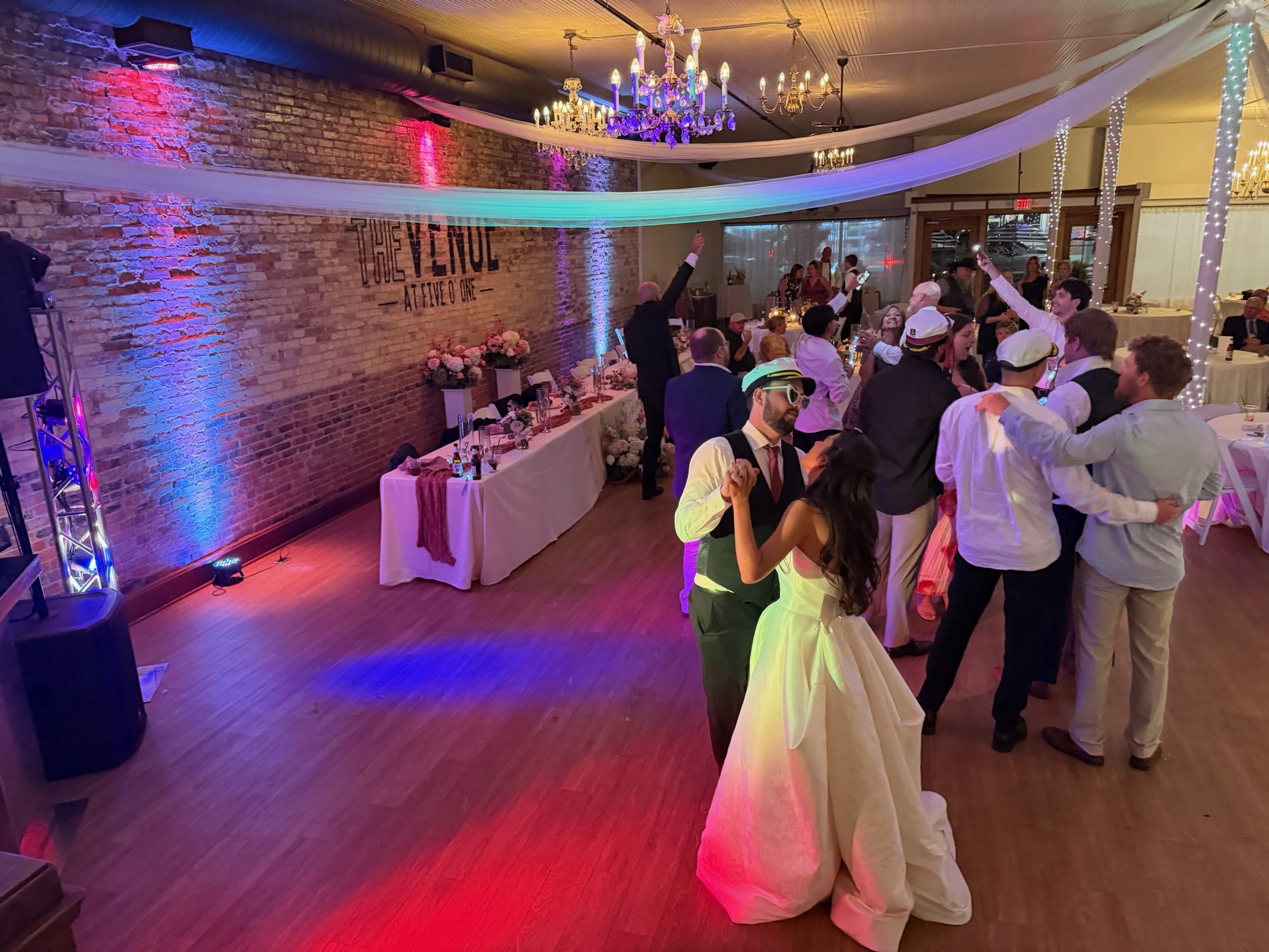 A bride and groom dancing with guests beneath colorful reception lighting